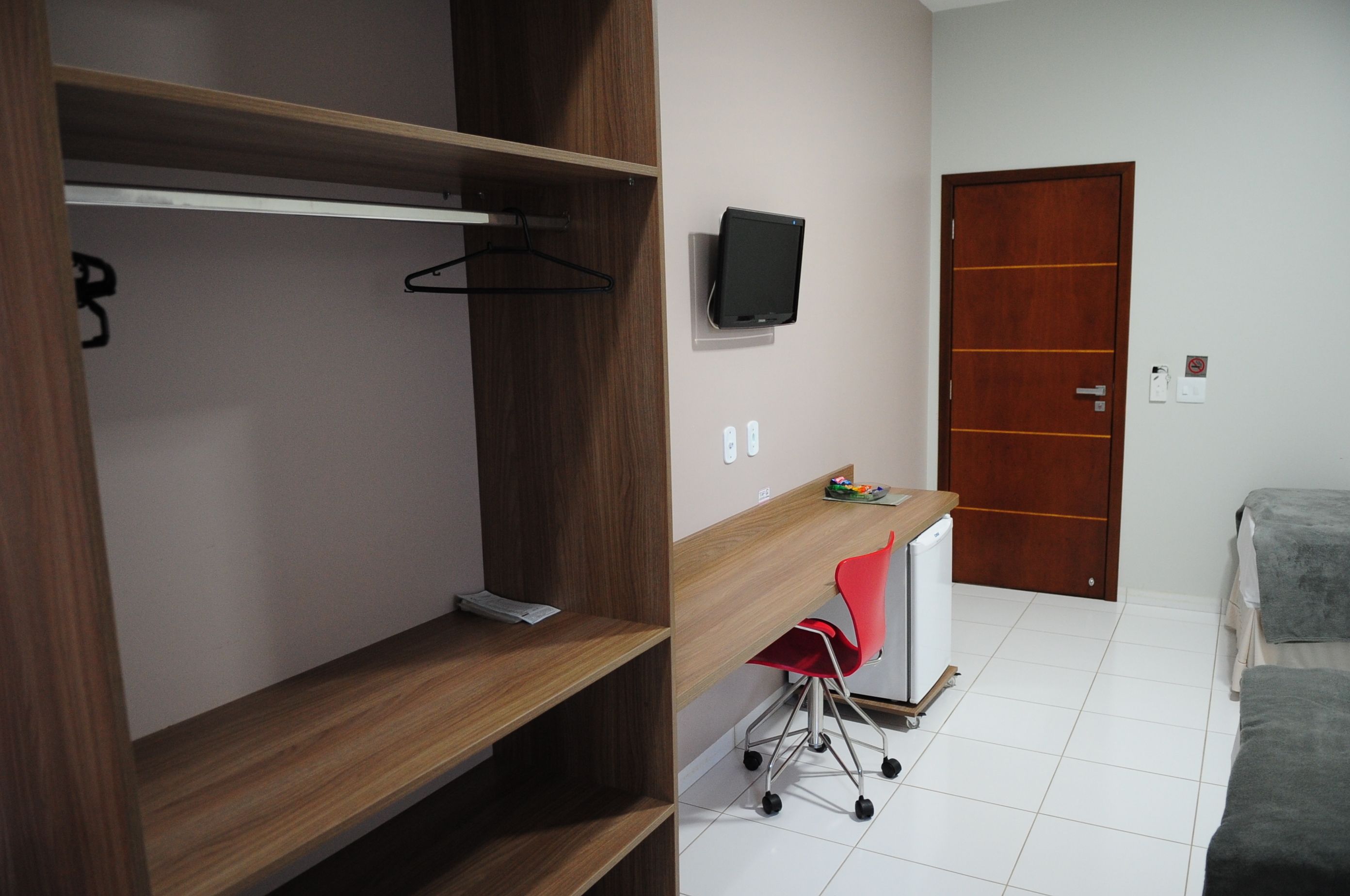 A room with a wooden wardrobe, a desk with a chair, a television on the wall, and a door.