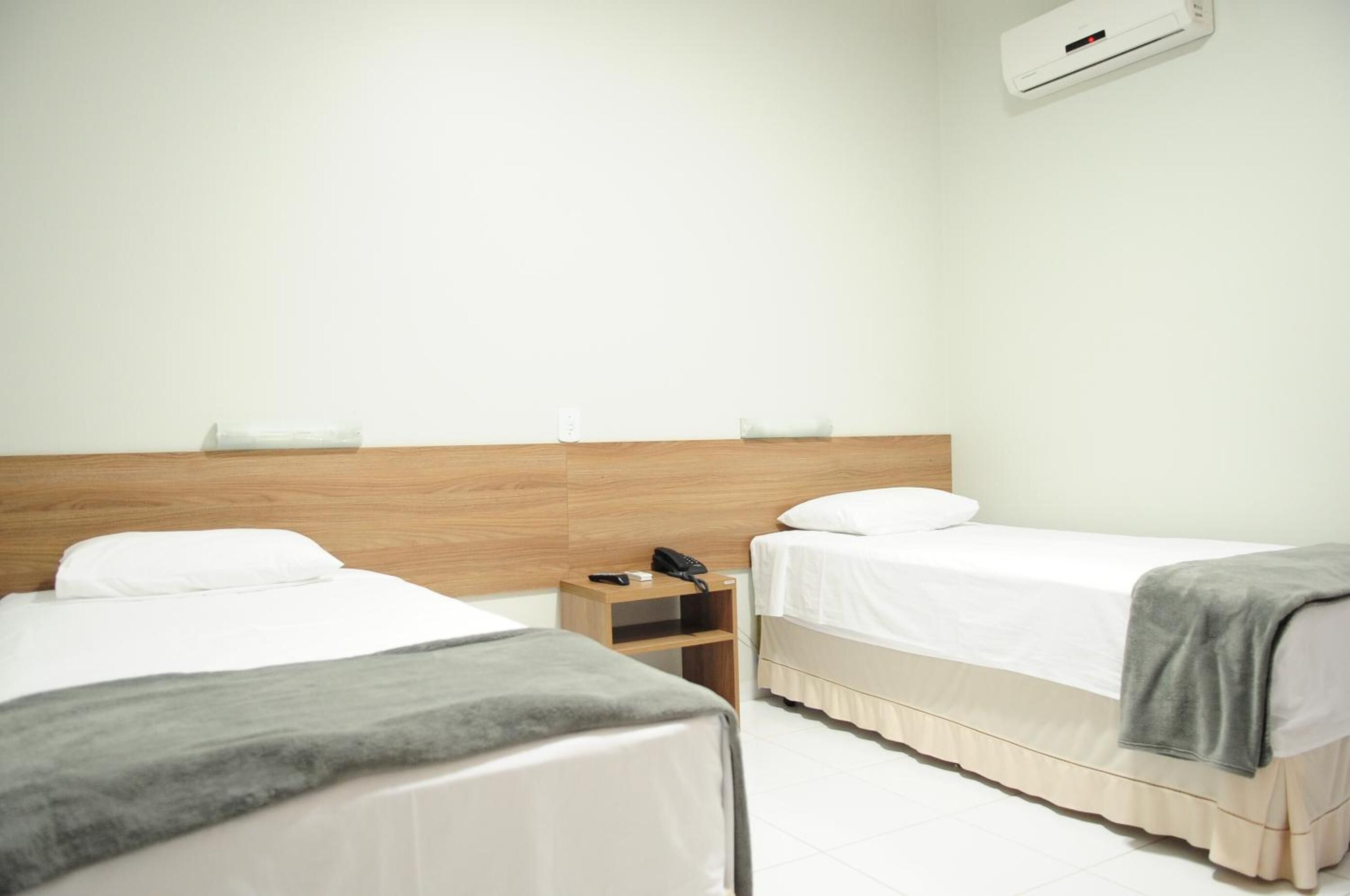 A hotel room with two beds, a wooden headboard, a bedside table with a phone, and a wall-mounted air conditioner.