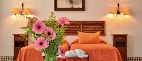 Habitación de hotel con una cama, dos mesitas de noche, un jarrón de flores rosadas y un cuadro en la pared.