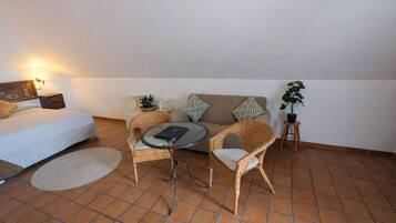 Une pièce comprenant un lit, un canapé, une table ronde, deux chaises en osier, une petite plante en pot et un luminaire fixé au mur.