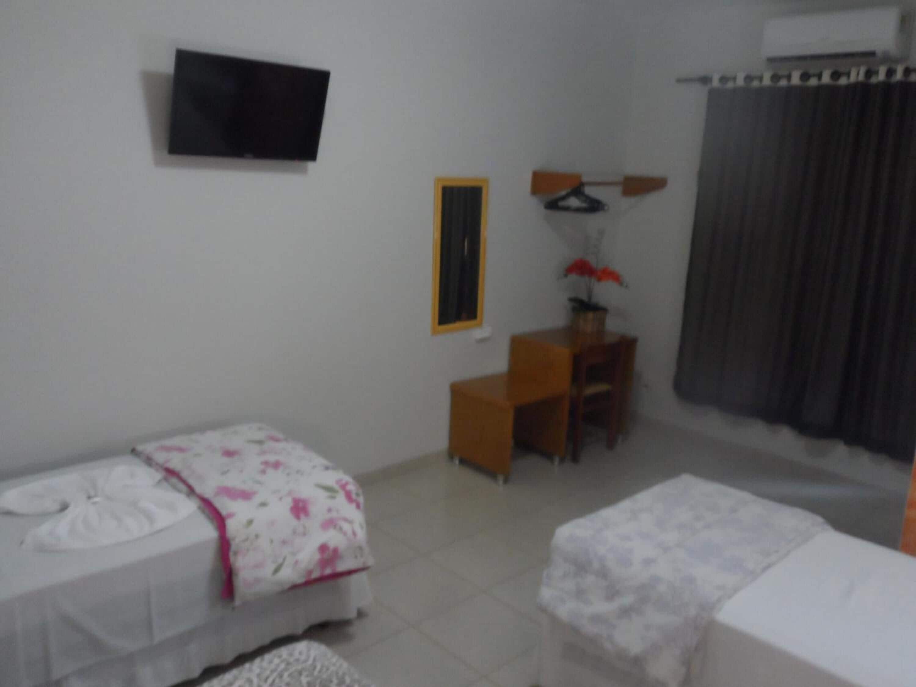 A small, well-lit room with a bed, a desk, a chair, and a TV mounted on the wall.