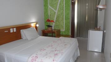 A hotel room with a bed, a small table with a plant, a desk, a chair, a refrigerator, and a curtain.