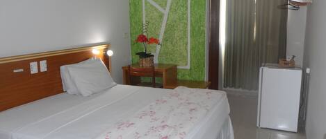 Una habitación de hotel con cama, una mesita con una planta, un escritorio, una silla, un refrigerador y una cortina.