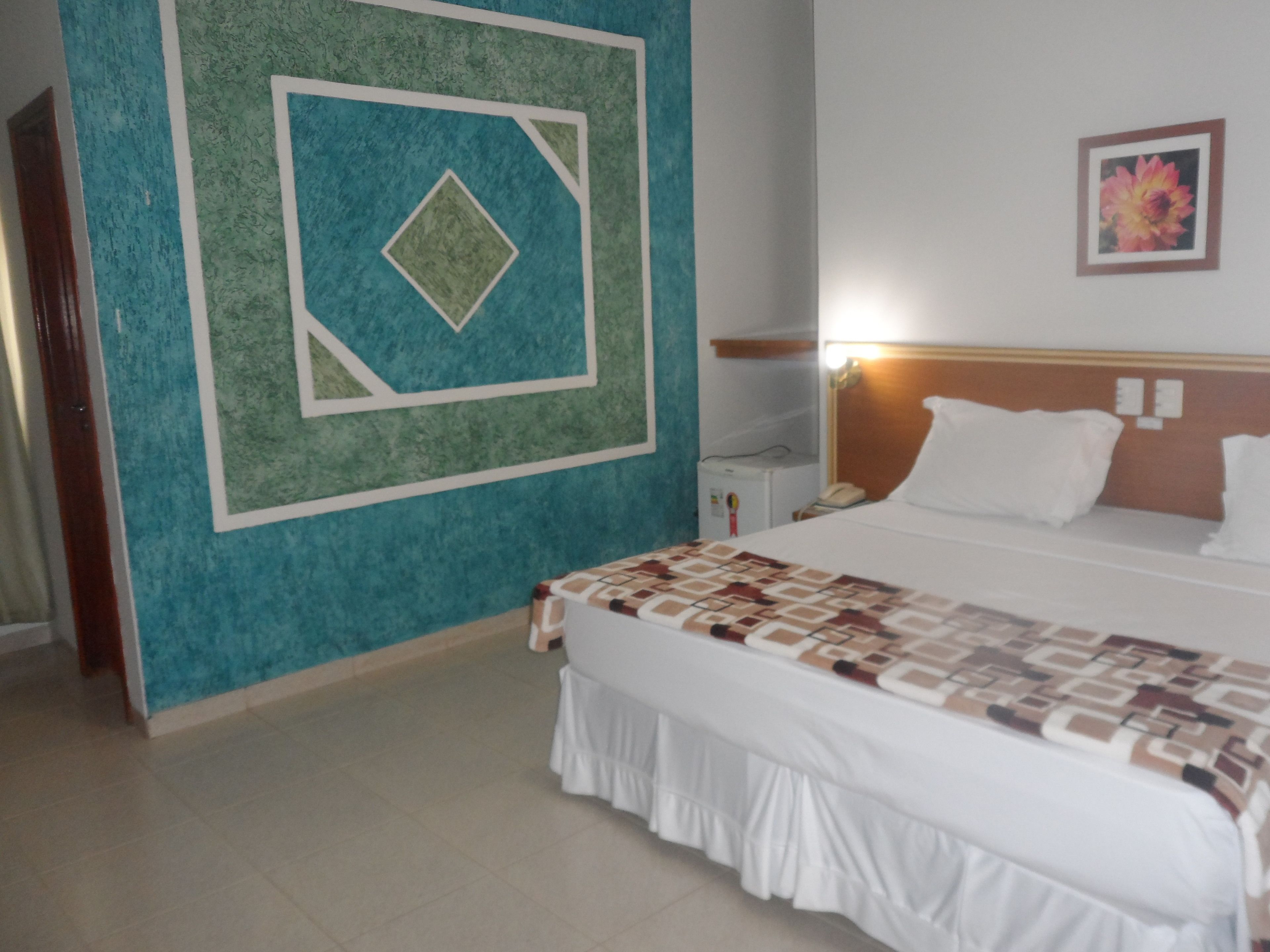 Quarto de hotel com uma cama, mesa de cabeceira, relógio de parede e um quadro na parede.