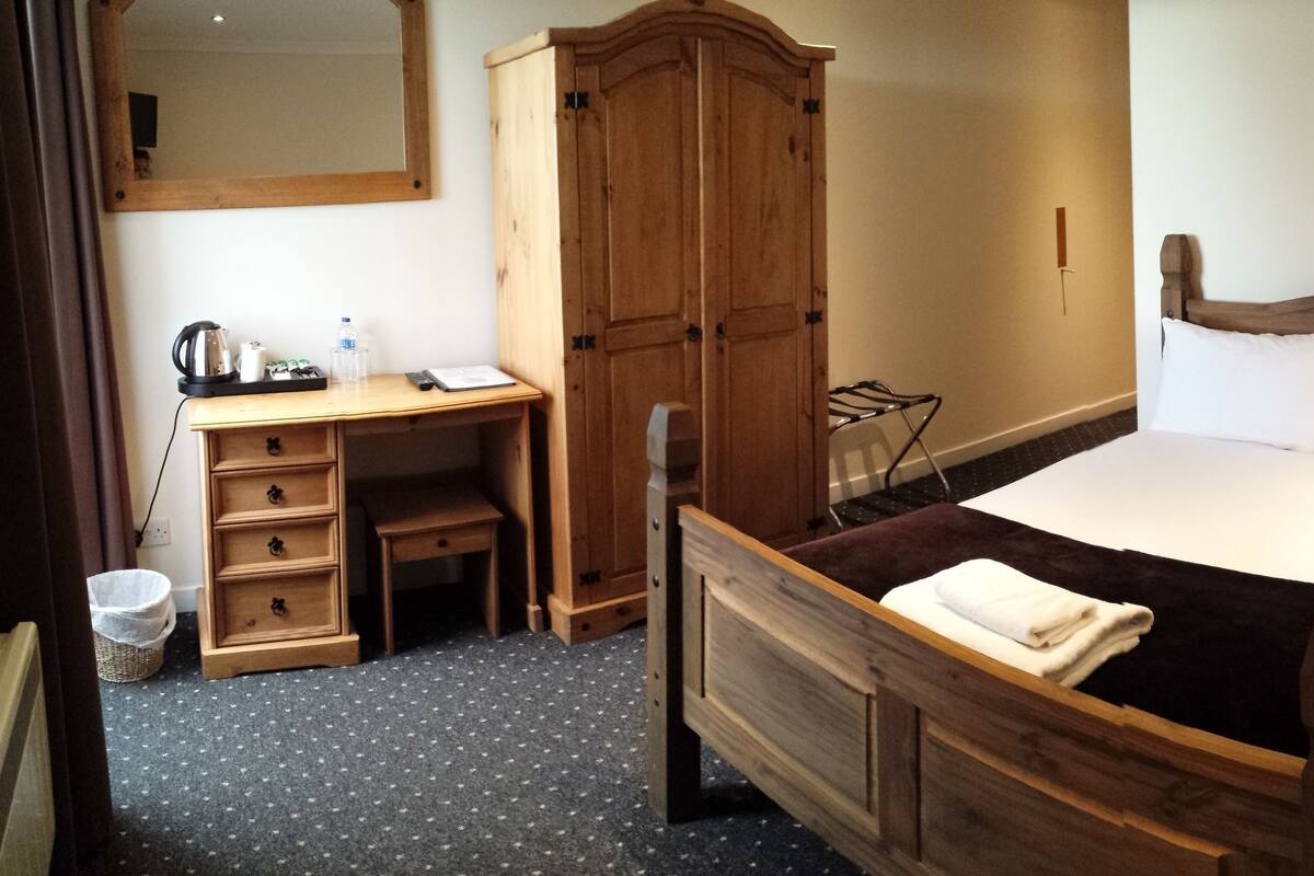 A wooden bed with a white pillow and a blanket, a wooden dresser, a wooden wardrobe, a wooden nightstand with a lamp, and a mirror on the wall.