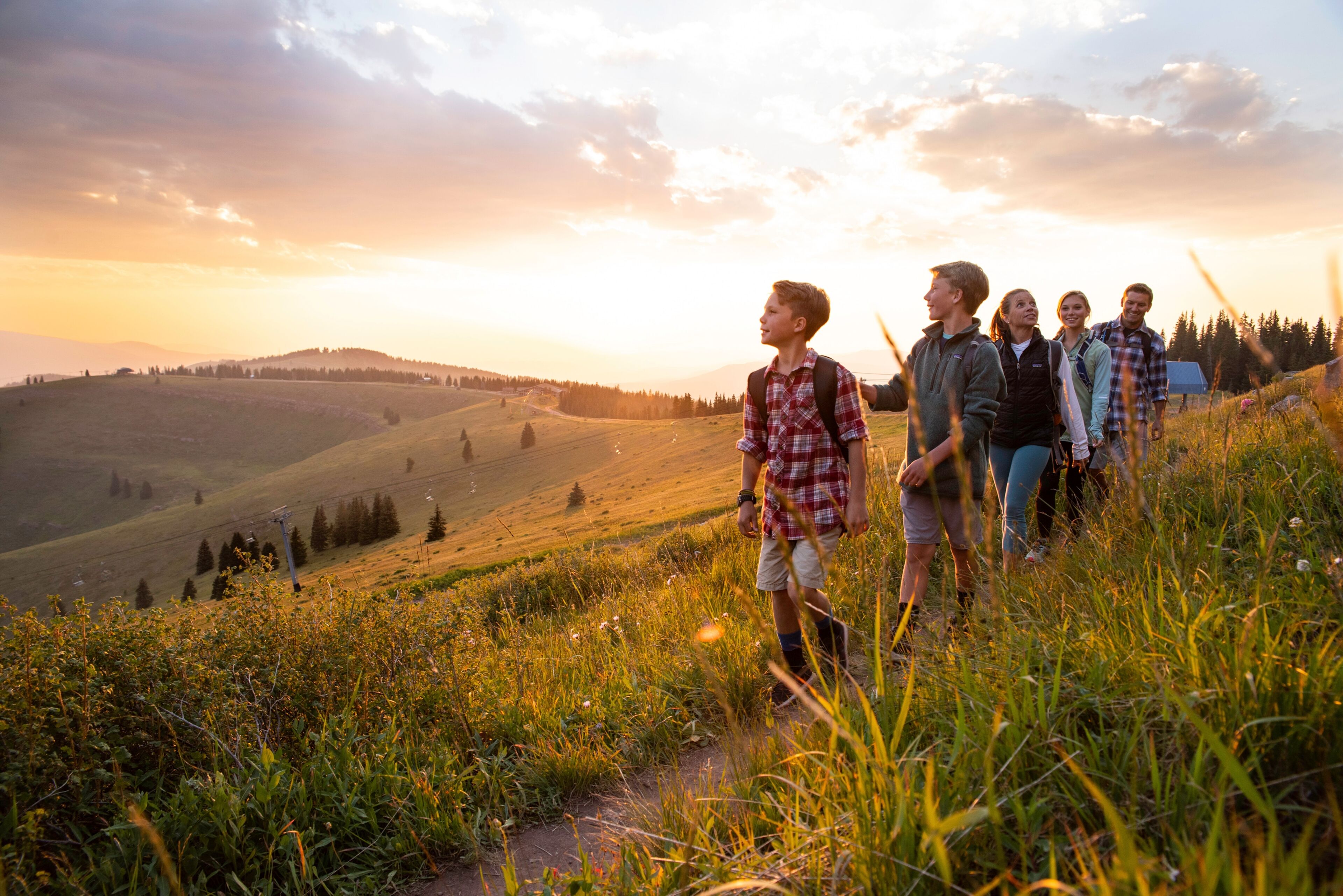 A group of people hiking on a grassy hillside with a scenic view of rolling hills and a sunset in the background.
