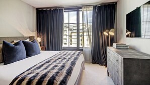 Habitación de hotel moderna con una cama grande, un aparador, un escritorio con lámpara y un amplio ventanal con cortinas.
