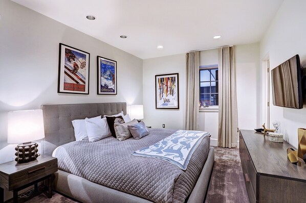 A modern hotel room with a large bed, a bedside table with a lamp, a dresser, and a TV mounted on the wall.