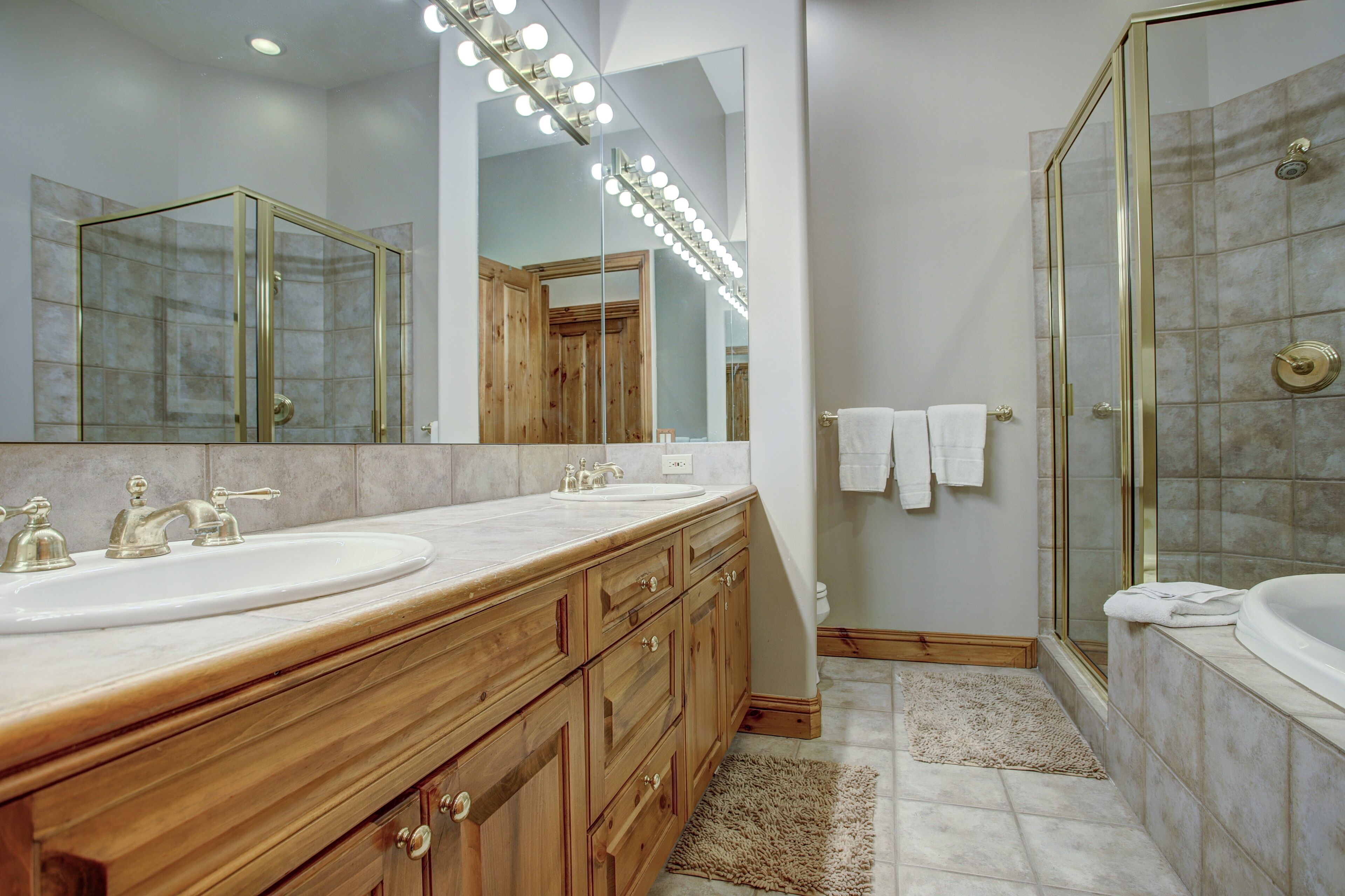 A bathroom with two sinks, a mirror with lights, a shower, and wooden cabinets.