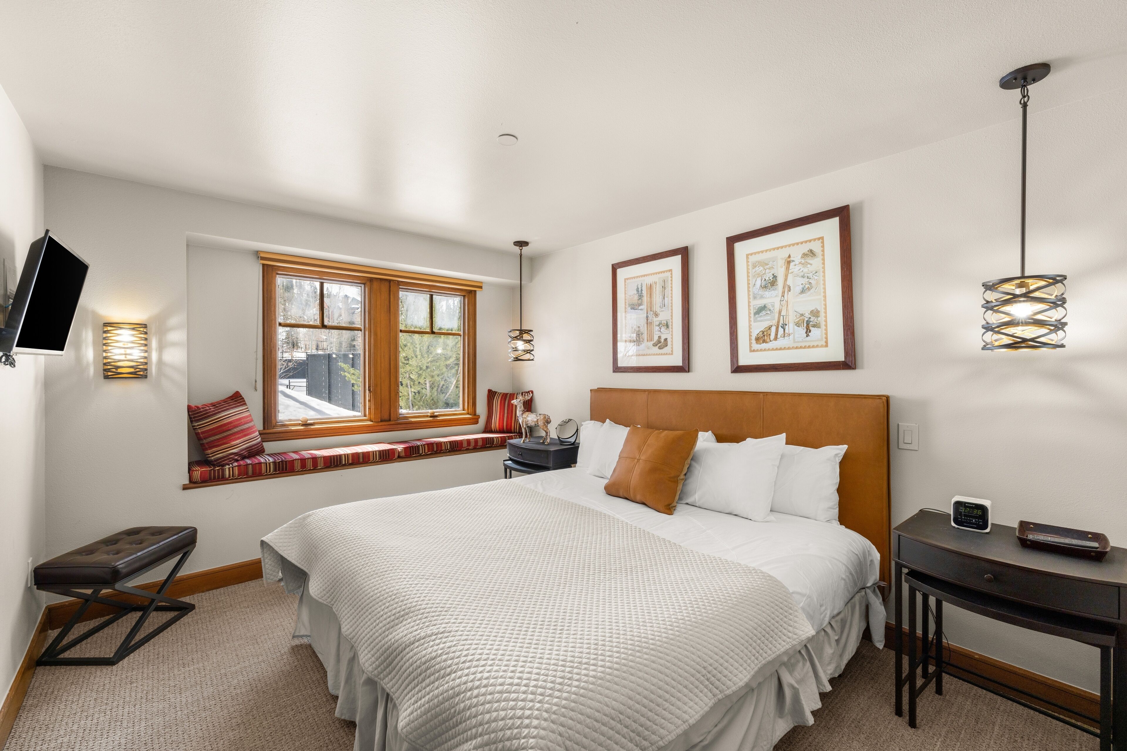 A bedroom with a bed, bedside table, lamp, window, artwork, and a TV mounted on the wall.