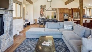 Ein geräumiges Wohnzimmer mit Kamin aus Stein, einem großen grauen Ecksofa, einem Essbereich mit Holztisch und einer Küche mit Holzschränken.