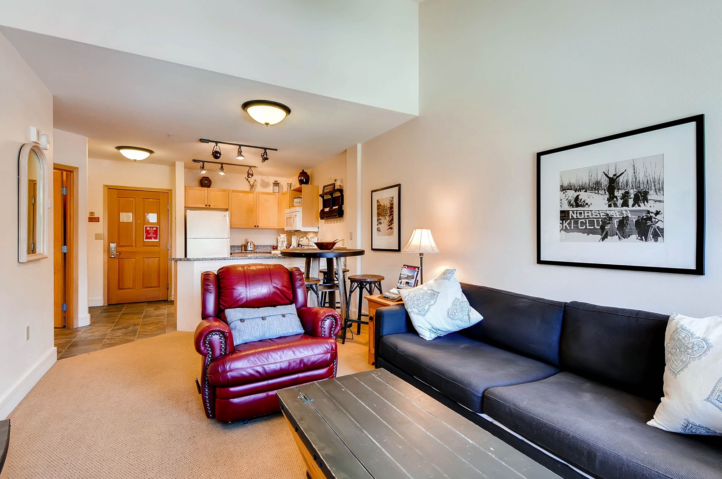A living room with a red leather chair, a black sofa, and a coffee table. There is a kitchen in the background with wooden cabinets and a refrigerator.