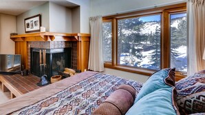 A cozy bedroom with a fireplace, a large window with a mountain view, and a bed with patterned bedding. - Lenawee 1747 (Keystone)
