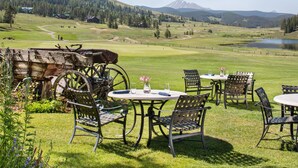 An outdoor seating area with metal chairs and tables, a rustic wooden cart, and a scenic view of a golf course and mountains. - Lenawee 1747 (Keystone)