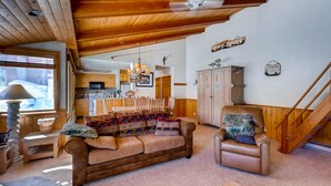 Un salon chaleureux avec un plafond en bois, un canapé, un fauteuil et un coin repas agrémenté d’un lustre.
