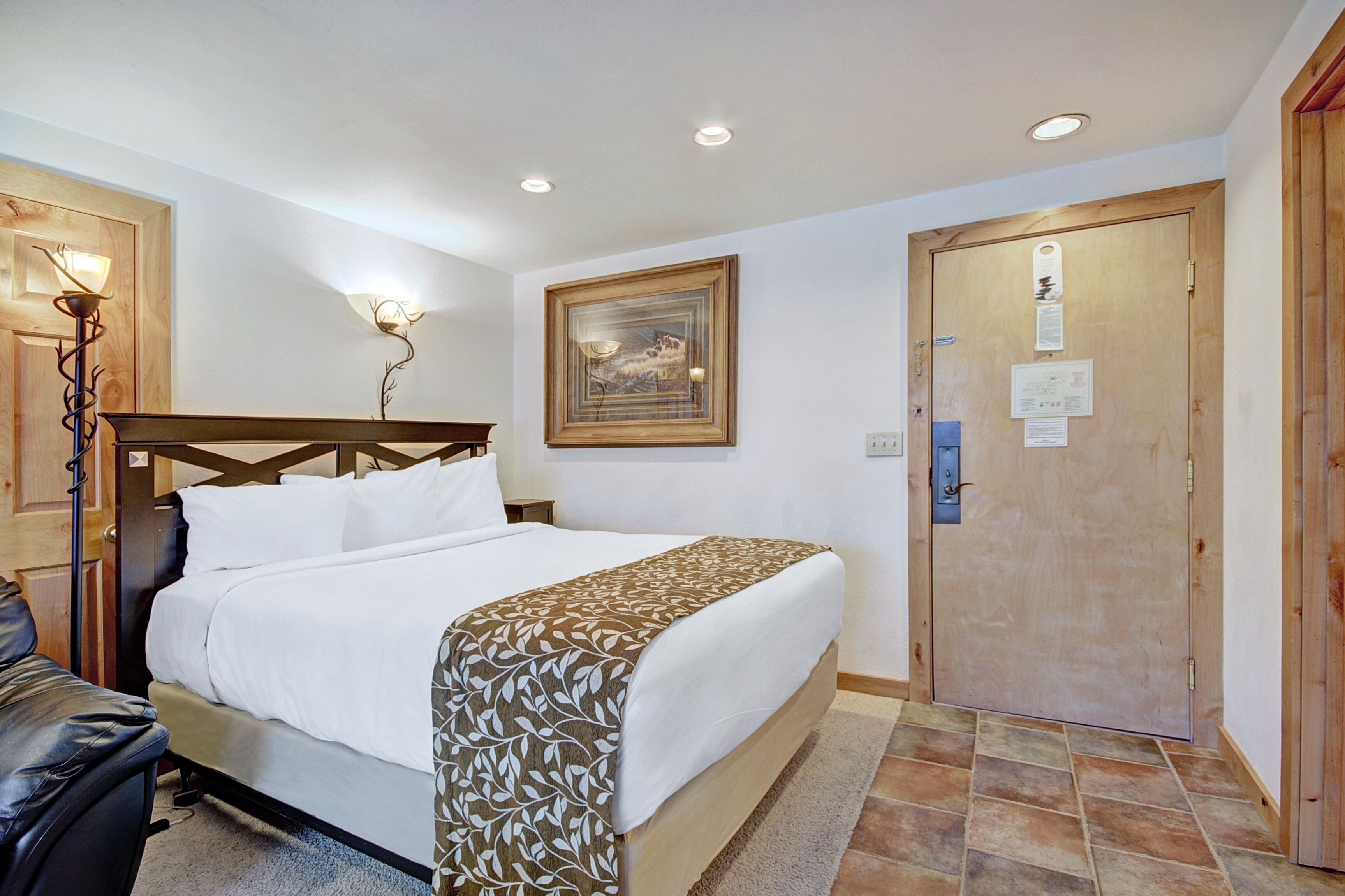 Une chambre d’hôtel avec un lit, une tête de lit en bois, une œuvre d’art accrochée au mur, une porte avec une serrure et un luminaire.