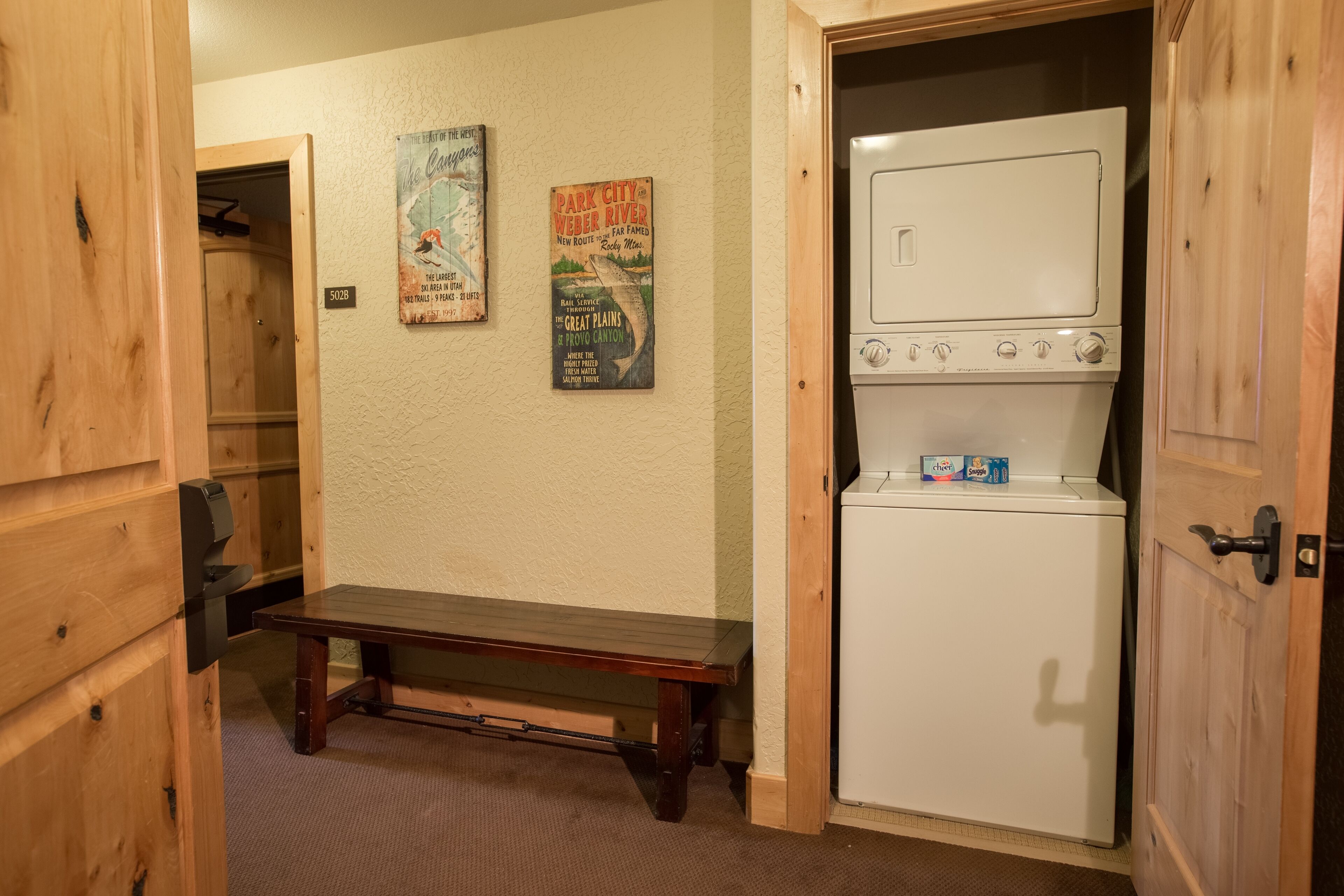 A compact room with a wooden bench, a white washer-dryer unit, and two vintage posters on the wall.