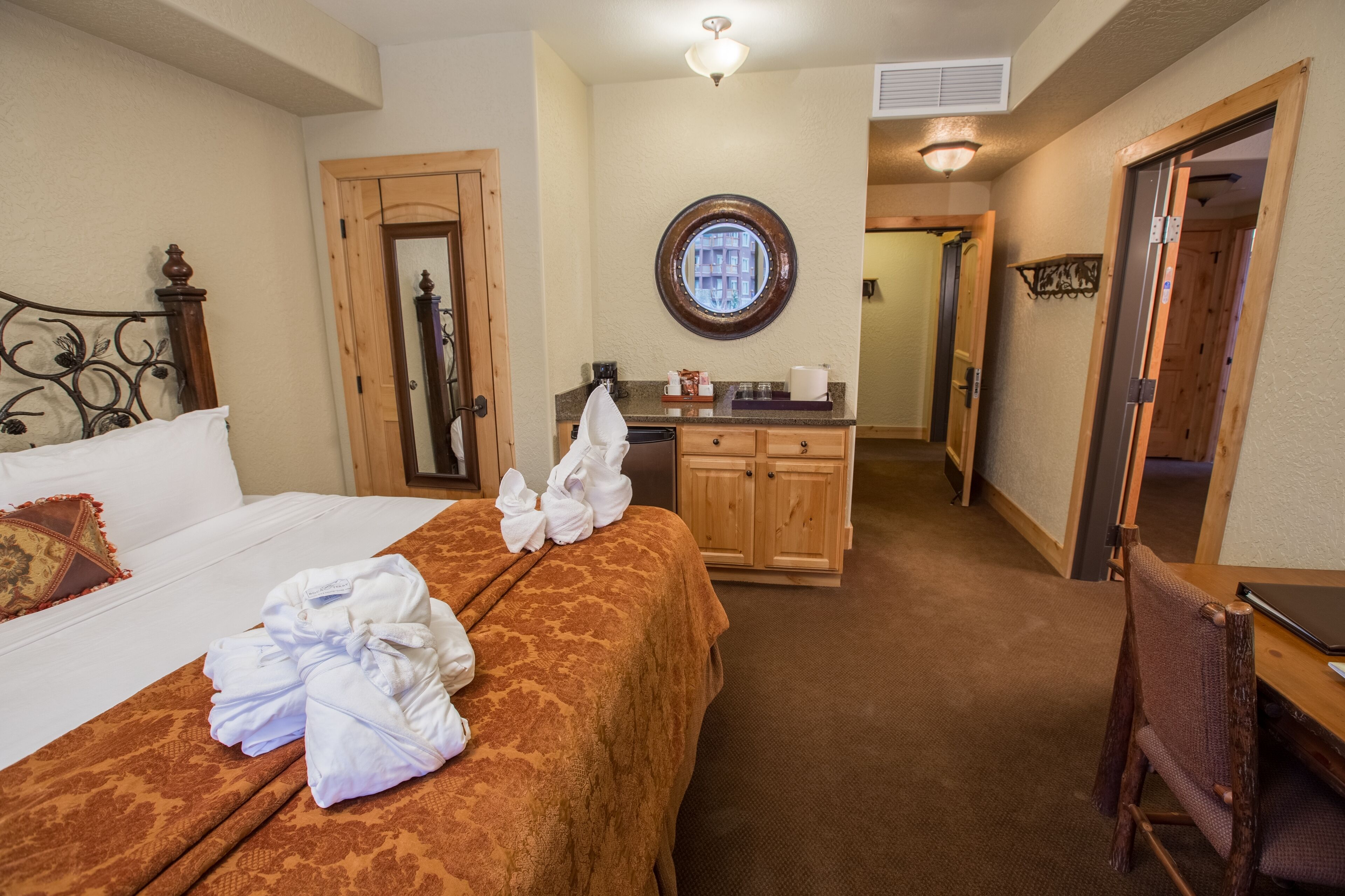 Une chambre d’hôtel avec un lit, une chaise en bois, un bureau, un miroir et une salle de bain visible à travers une porte ouverte.