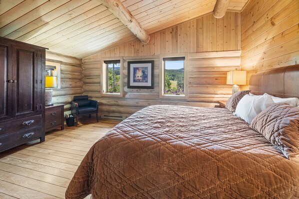 A log cabin bedroom with a large bed, a dresser, a desk, and two windows with framed pictures.
