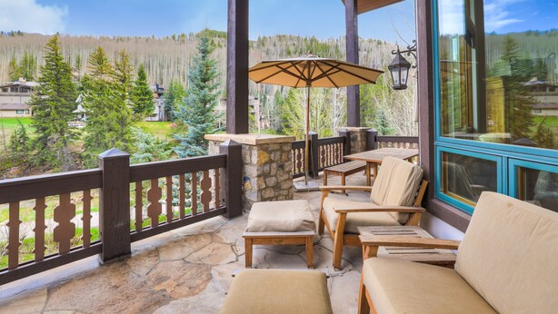 A covered outdoor seating area with stone flooring, wooden furniture, and a view of a forested landscape. - Refined 5 BR Private Home with Private Hot Tub by RedAwning (Vail)