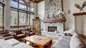 A spacious living room with a stone fireplace, leather furniture, and large windows offering a view of the forest. - Refined 5 BR Private Home with Private Hot Tub by RedAwning (Vail)