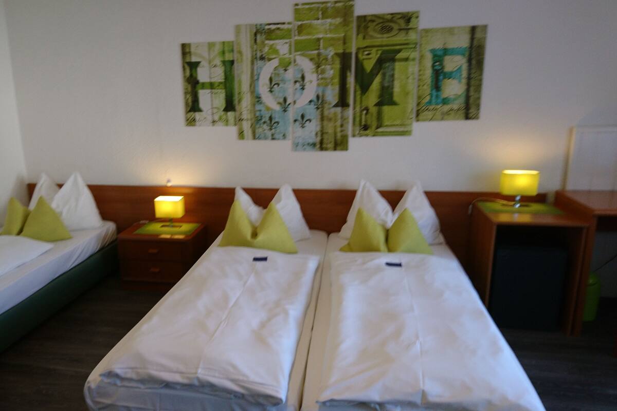 Quarto de hotel com duas camas de solteiro, uma cama de casal e uma mesa de cabeceira com abajur.