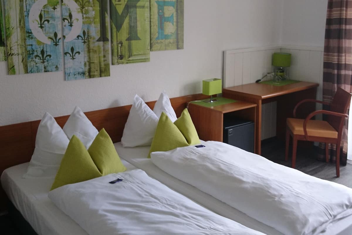 Uma cama de casal com roupa de cama branca e verde, uma escrivaninha com abajur verde e uma cadeira.