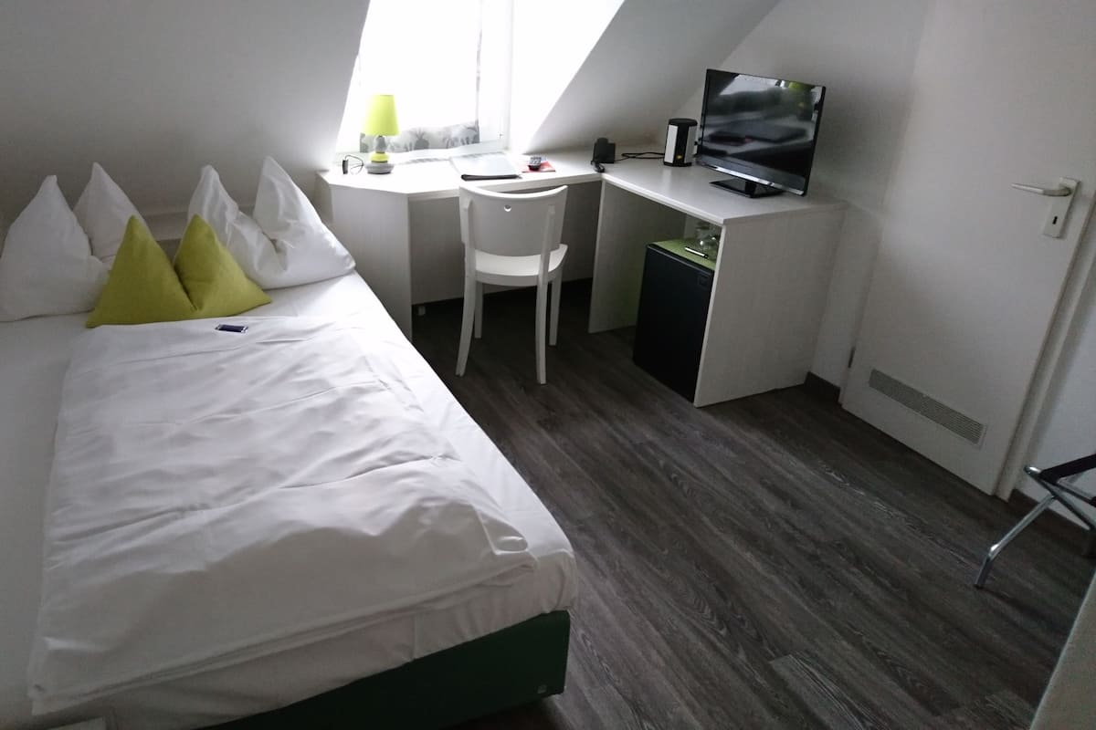 Um quarto pequeno e bem arrumado, com uma cama, uma escrivaninha, uma cadeira e uma televisão.