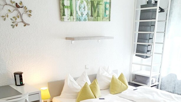 Ein ordentlich eingerichtetes Schlafzimmer mit Bett, Schreibtisch und Stuhl. Über dem Bett befindet sich eine Wanddekoration und rechts eine Regalwand.