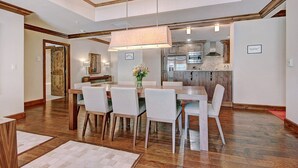 A spacious dining area with a long table and multiple chairs, a hanging light fixture, and a view into the kitchen area.
