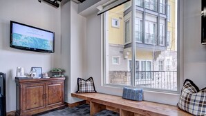 A cozy seating area with a wooden bench and cushions, a television mounted on the wall, and a window offering a view of the city.