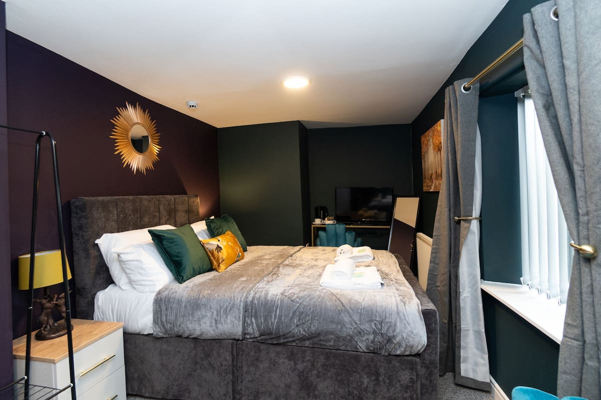 A modern bedroom with a large bed, a bedside table with a lamp, a wall-mounted mirror, and a window with curtains.