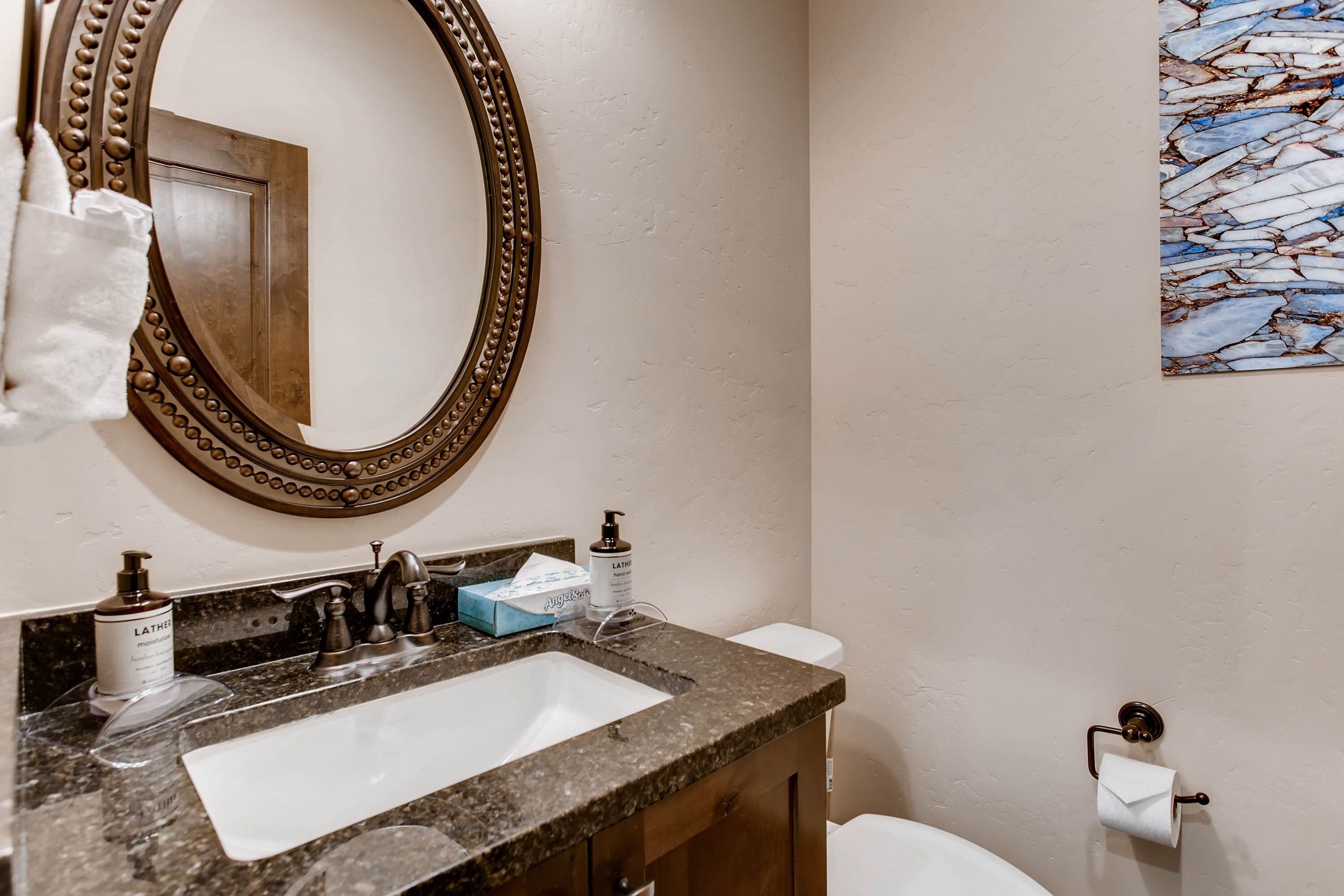 A bathroom with a granite countertop, a white sink, a mirror with a decorative frame, toiletries, and a toilet.