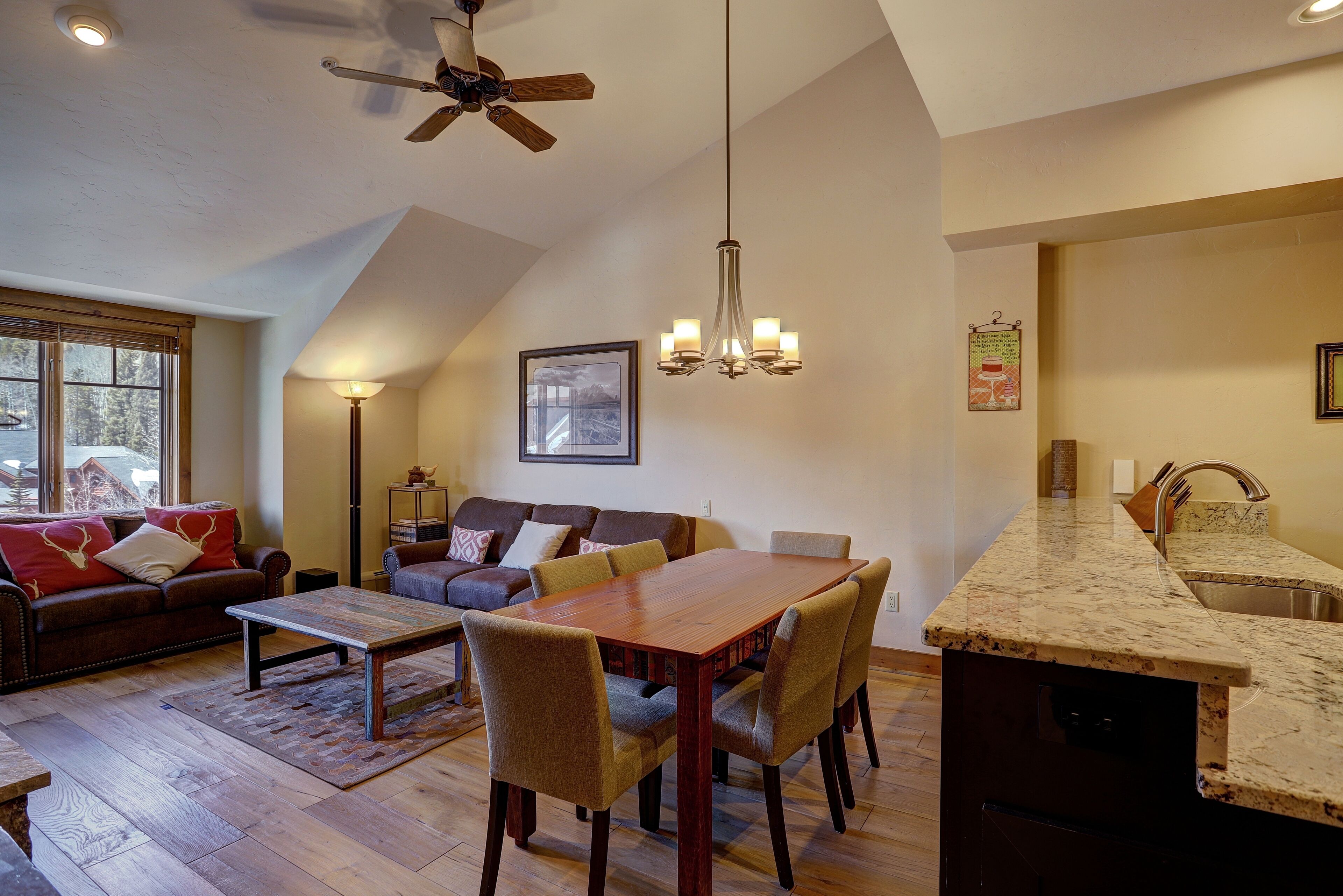 A spacious living room with a wooden dining table, comfortable seating, a ceiling fan, and a kitchen area with a granite countertop.