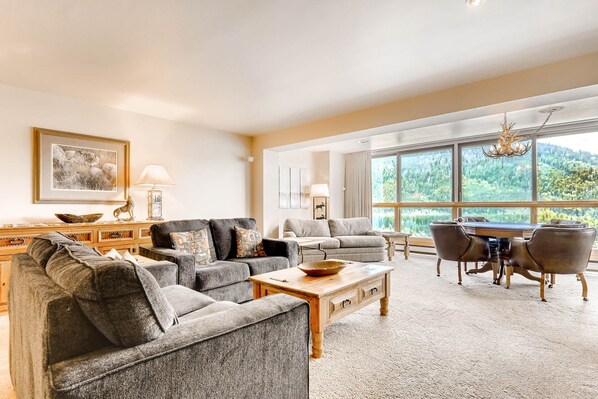 A spacious living room with a large window, a wooden coffee table, and a dining area with a chandelier. - Spacious Penthouse 3br ~no Cleaning Fee~kids Ski Free! 3 Bedroom Condo by RedAwning (Keystone)