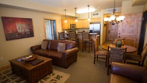 A living room with a brown leather sofa, a wooden coffee table, and a patterned rug. There is a dining area with a wooden table and chairs, and a kitchen with wooden cabinets and a refrigerator. - Valley Views with Loaded Bedrooms by RedAwning (Park City)