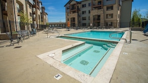 A pool area with two swimming pools, lounge chairs, and a building with multiple balconies in the background.