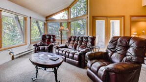 Ein geräumiges Wohnzimmer mit Ledersofas, einem runden Couchtisch und großen Fenstern mit Blick auf Bäume.