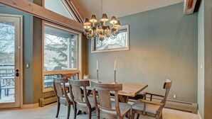 A dining area with a wooden table and chairs, a chandelier, and a large window with a view of trees. - Slopeside Living - 3br 4ba At Pines Lodge 3 Bedroom Condo by RedAwning (Avon)