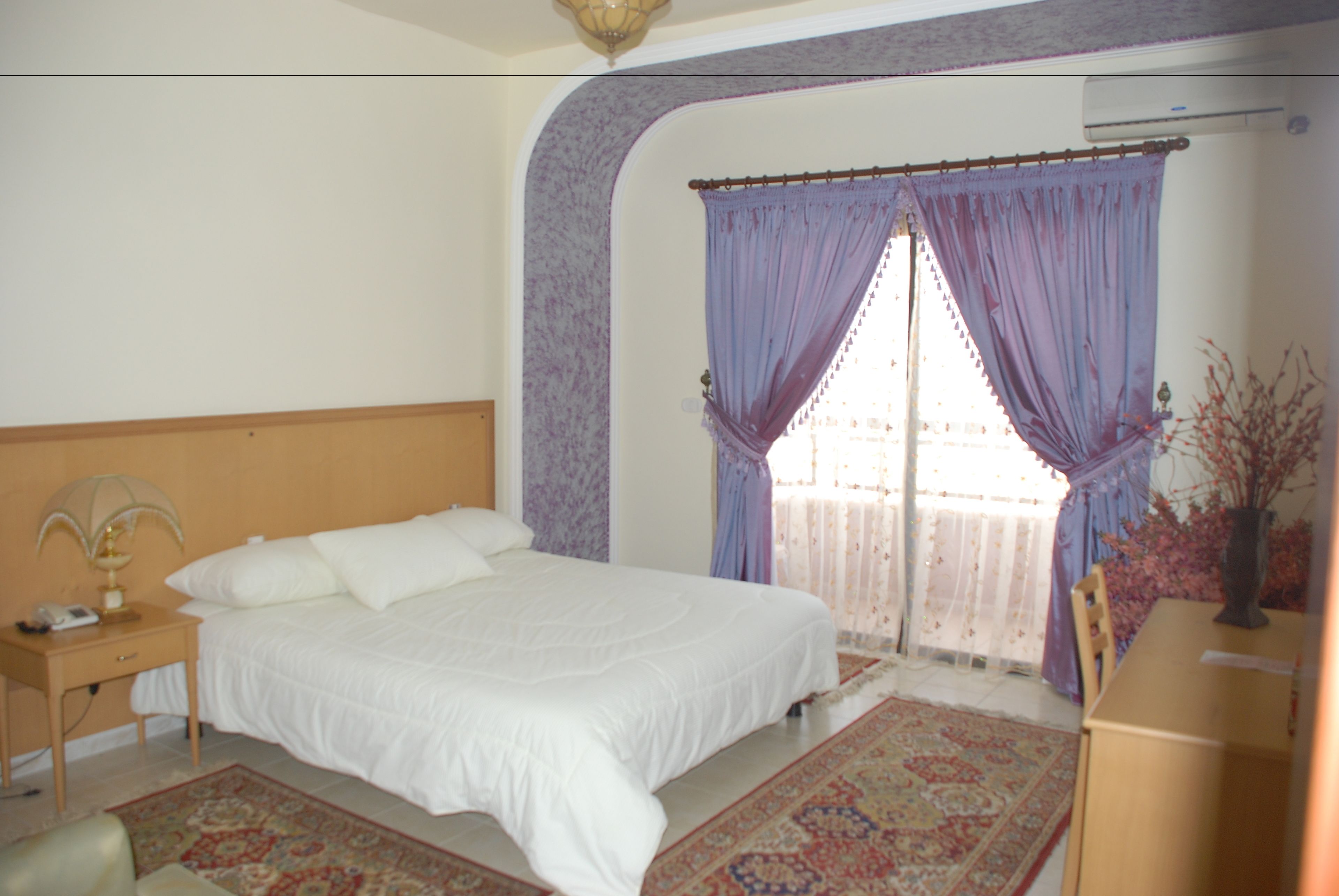 A bedroom with a large bed, a nightstand, a desk, and a window with purple curtains.