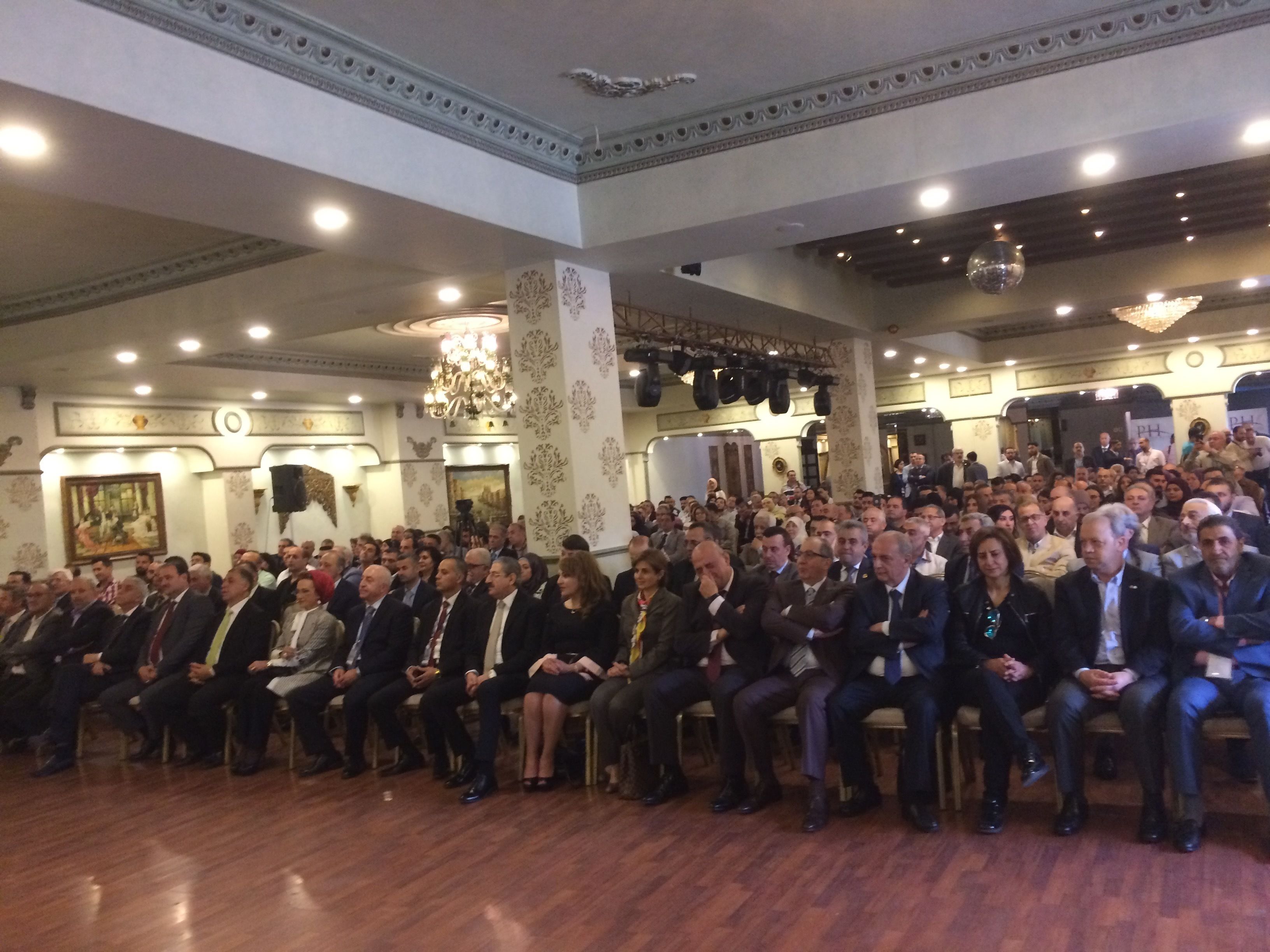 A formal indoor gathering with attendees seated in rows, a chandelier, and ornate wall decorations.