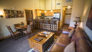 Ein Wohnzimmer mit braunem Ledersofa, einem Holztisch und einem Essbereich mit Holztisch und Stühlen. Es gibt eine Küche mit Holzschränken und einem Kühlschrank.