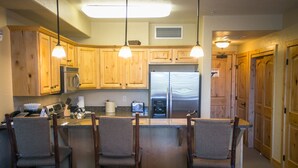 A kitchen with wooden cabinets, a refrigerator, a microwave, and a coffee maker. There are four bar stools and a countertop with a kettle. - Stay in Comfort while Enjoying Valley Views by RedAwning (Park City)