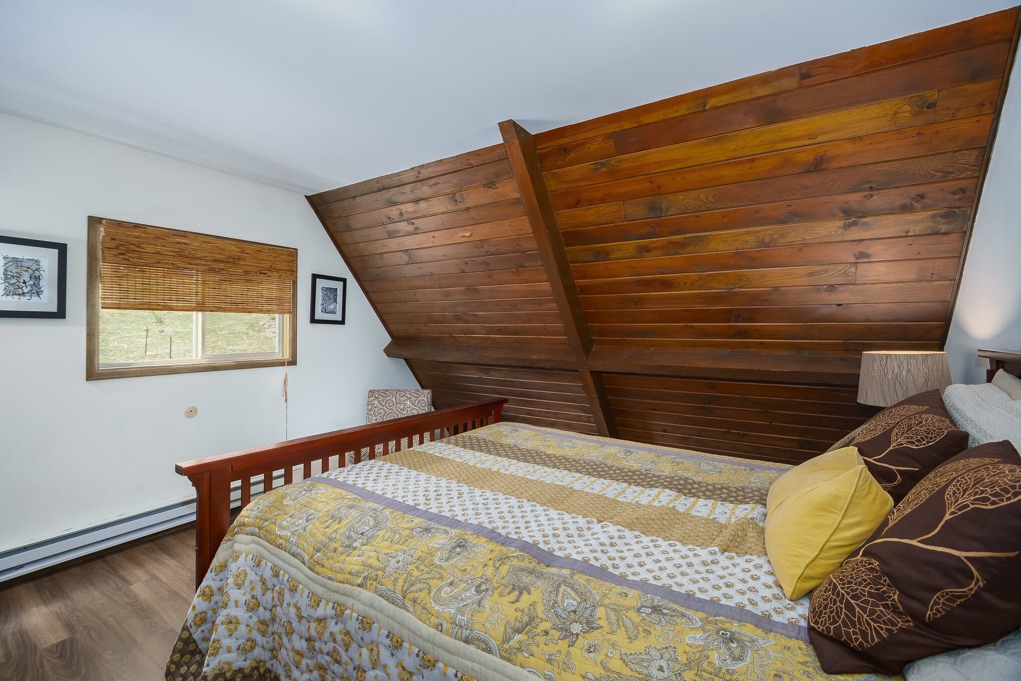 A bedroom with a sloped ceiling, a bed with patterned bedding, a window with blinds, and a wooden headboard.