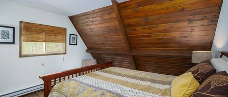 A bedroom with a sloped ceiling, a bed with patterned bedding, a window with blinds, and a wooden headboard.
