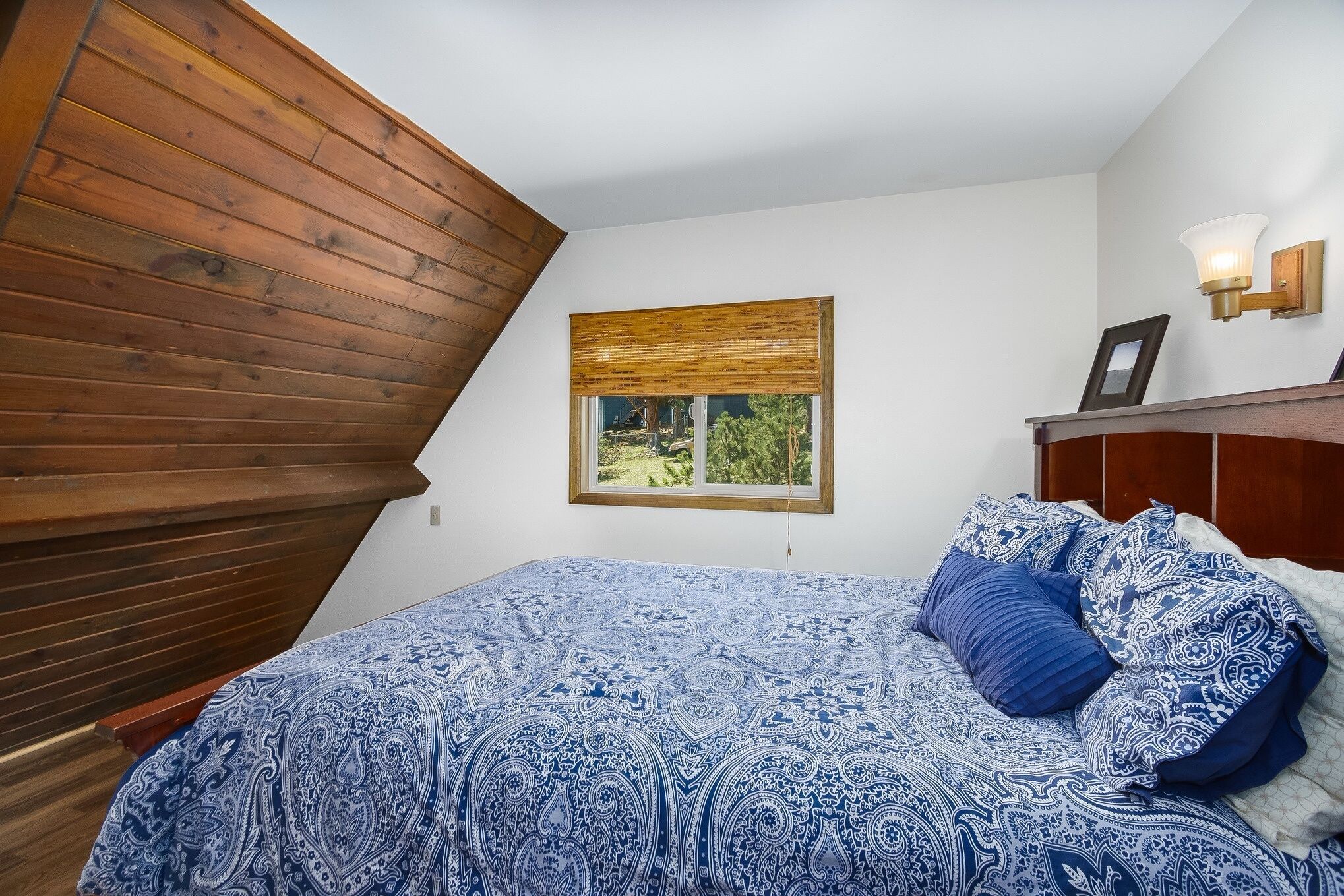 Une chambre avec un plafond en pente, un lit recouvert d’une couvre-lit à motifs bleus, une fenêtre donnant sur la verdure et un luminaire fixé au mur.
