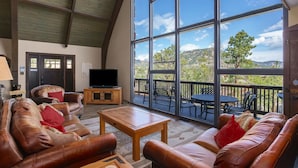 A spacious living area with leather sofas, a wooden coffee table, and a large window offering a view of a deck and mountains. - Sky View In The Rockies -- Ev #3473 3 Bedroom Home by RedAwning (Estes Park)