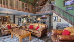 A spacious living room with leather sofas, a wooden coffee table, and a stone fireplace. A wooden staircase with a railing leads to an upper level. - Sky View In The Rockies -- Ev #3473 3 Bedroom Home by RedAwning (Estes Park)