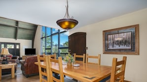 A dining area with a wooden table and chairs, a chandelier, a painting, and a view through large windows. - Sky View In The Rockies -- Ev #3473 3 Bedroom Home by RedAwning (Estes Park)
