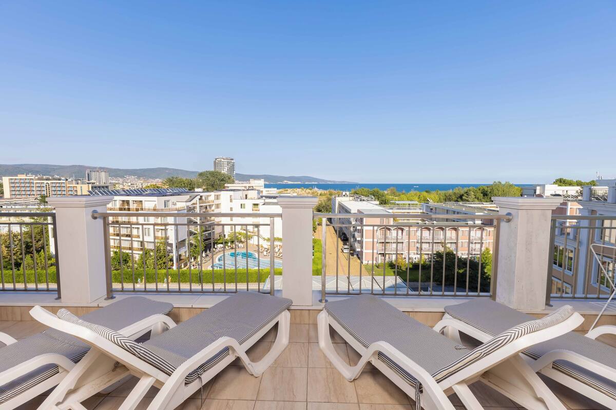 Un balcón con sillones de sol, vista a una piscina y edificios circundantes.